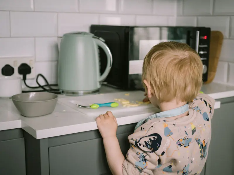 A toddler reaching for food on the kitchen counter next to a kettle and microwave