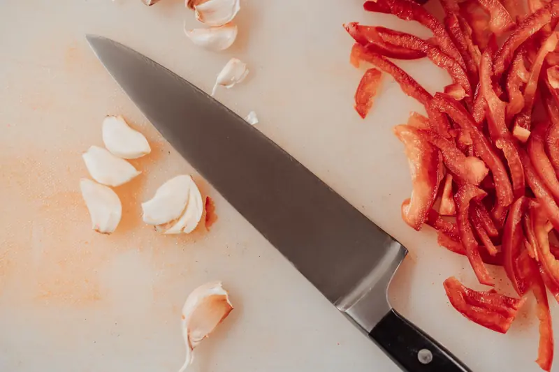 Top view of a kitchen knife with sliced red peppers and garlic on a chopping board