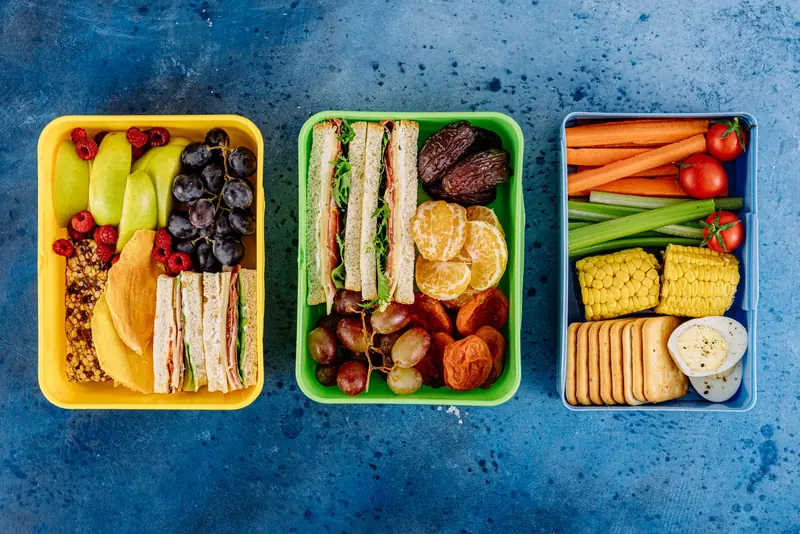 Three vibrant lunch boxes showcasing fruits, sandwiches, and vegetables on a blue textured background